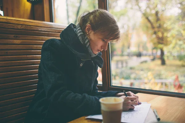 Cup of coffee on the foreground with elegant young woman at the coffee shop wooden table, work break of business people, flare sun light . Cold autumn, the female draws or writes in diary