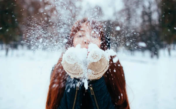 Inverno. Mulher de cabelo ruivo usando abafadores de ouvido soprando na neve nas mãos — Fotografia de Stock