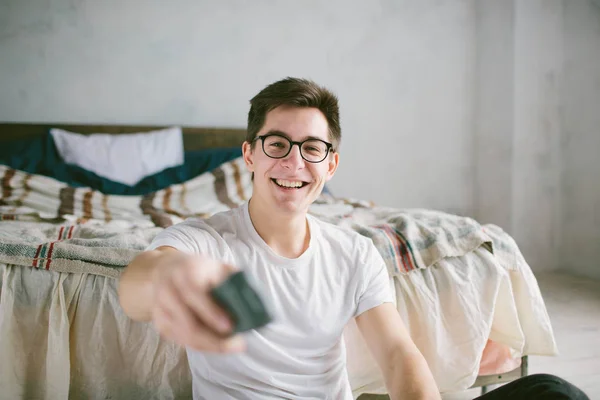 Guy using remote control while sitting on the bed in home. Happy attractive young man watching tv . Human emotions on her face. — Stock Photo, Image