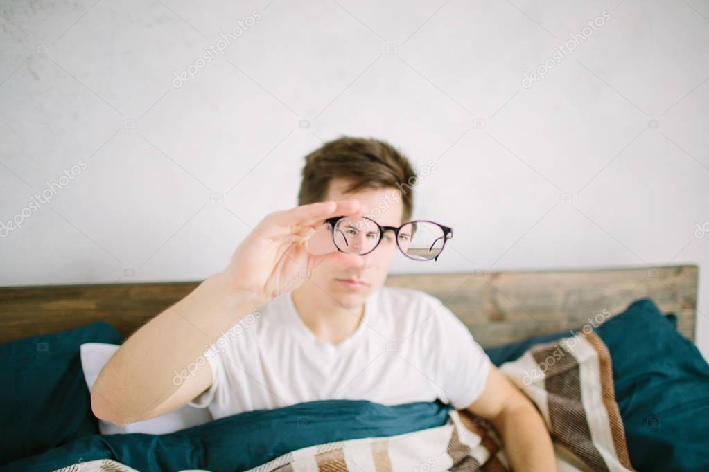 Closeup portrait of young man with glasses. He has eyesight problems and is squinting his eyes a little bit. Handsome guy is holding his eyeglasses right in front of camera with one hand