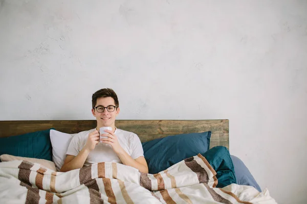 这是一个美妙的早晨。一个年轻人在床上醒来。男人躺在床上，晨光下喝咖啡. — 图库照片