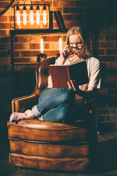 Girl in a cozy dark room reading a book with glasses. Blonde and warm lamp light