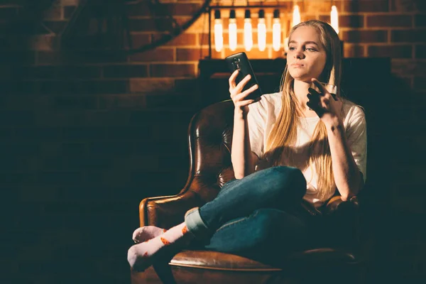 Mädchen in einem gemütlichen dunklen Raum hält ein Telefon in der Hand und durchsucht das Internet. Blondes und warmes Lampenlicht — Stockfoto