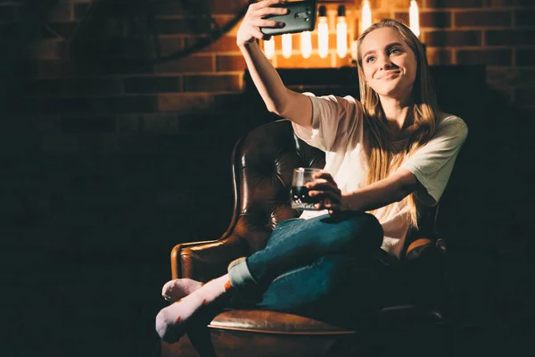 Flicka i ett mysigt mörkt rum med en telefon. Blond och varm lampa ljus — Stockfoto