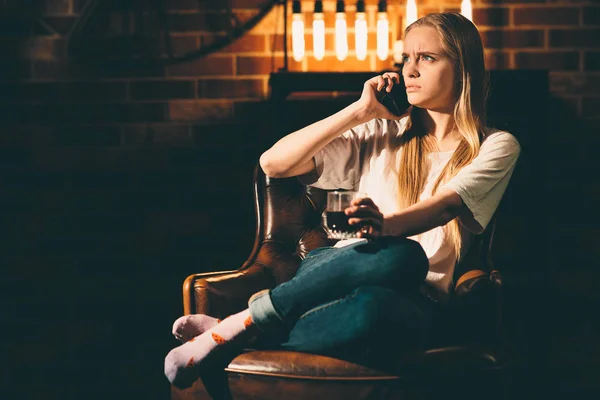 Menina em um quarto escuro aconchegante está sentado em uma cadeira e falando ao telefone. Luz de lâmpada loira e quente — Fotografia de Stock
