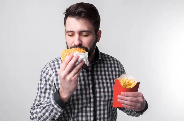 Un hombre sosteniendo un pedazo de hamburguesa y papas fritas. Estudiante come comida rápida. comida no útil. tipo muy hambriento — Foto de Stock