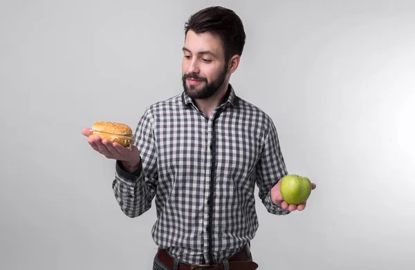 Homem barbudo em camisa quadriculada em um fundo claro segurando um hambúrguer e uma maçã. O tipo faz a escolha entre comida rápida e saudável. Saboroso ou útil O dilema — Fotografia de Stock