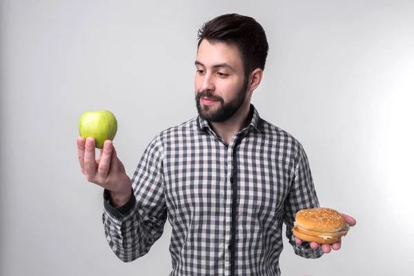 Bebaarde man in geruite overhemd op een lichte achtergrond holding een hamburger en een appel. Man maakt de keuze tussen snelle en gezonde voeding. Smakelijke of nuttig het dilemma — Stockfoto