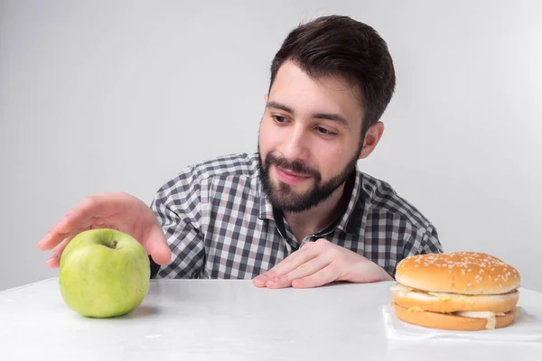 Bearded man in checkered shirt on a light background holding a hamburger and an apple. Guy makes the choice between fast and healthy food. Tasty or useful The dilemma — Stock Photo, Image