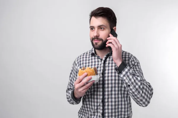 Hombre barbudo con camisa a cuadros sobre un fondo claro sosteniendo una hamburguesa y una manzana. Chico hace la elección entre la comida rápida y saludable. Sabroso o útil El dilema — Foto de Stock