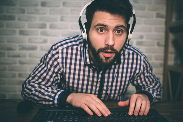 Bearded man playing at home and streaming. Funny guy is excited because of seen virtual video. He is wearing red headphones — Stock Photo, Image