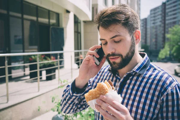 Handsome sporty bearded dark-haired man eating a hamburger and talking on the phone — Stock Photo, Image