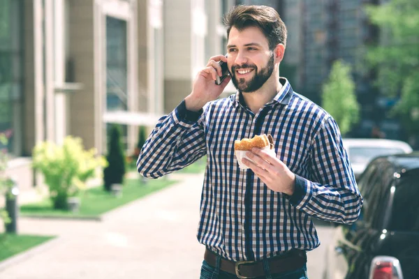 Handsome sporty bearded dark-haired man eating a hamburger and talking on the phone — Stock Photo, Image
