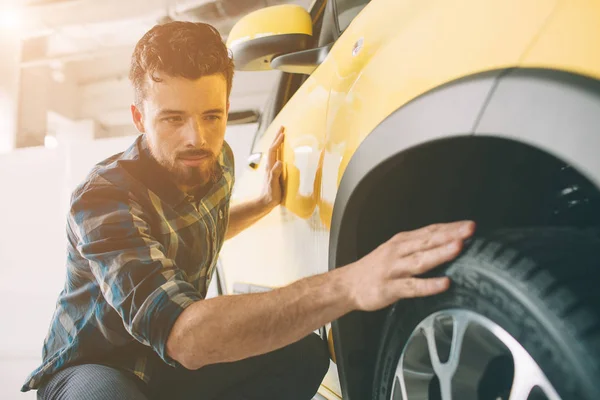 Líneas perfectas. El joven moreno barbudo examinando el coche en el concesionario y haciendo su elección. Retrato horizontal de un joven en el coche. Está pensando si debería comprarlo. — Foto de Stock