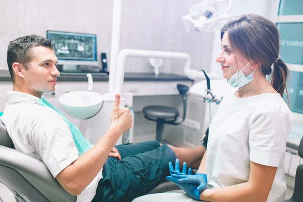 Portrait d'une dentiste et d'un jeune homme heureux dans un cabinet de dentiste. L'homme abandonne les pouces — Photo