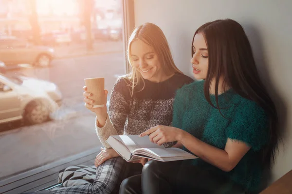 Két lány s szívódik fel az olvasó a könyvet, a szünetben a kávézóban. Aranyos szép fiatal nők könyvet olvas, és a kávéfogyasztás — Stock Fotó