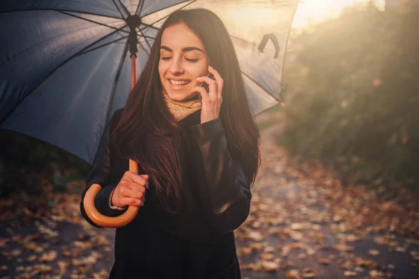在秋季使用智能手机的妇女。秋天女孩在太阳耀斑树叶的智能电话交谈。秋季色彩中的高加索森林模型肖像 — 图库照片