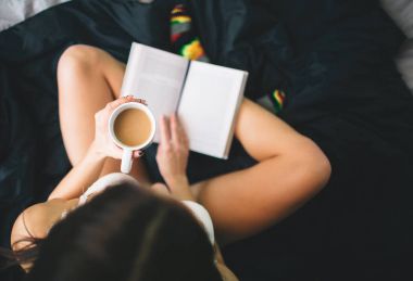 Kitap okuma ve yatakta kahve içme kız. Üstten Görünüm genç kadının siyah çarşaf üzerine bir kitap okumak.