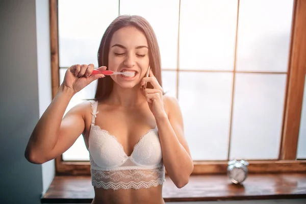 Портрет усміхненої свіжої брюнетки молода жінка в білій білизні розмовляє по телефону при чищенні зубів — стокове фото