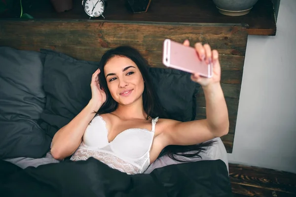 Érzéki fiatal nő, a fekete fehérnemű, hogy selfie egy okostelefon, és mosolyogva, miközben ágyon fehér fehérnemű. — Stock Fotó