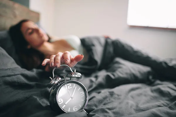 Wanita muda tidur dan jam alarm di kamar tidur di rumah. Gadis ketiduran di tempat tidur dan mencari jam alarm shock — Stok Foto