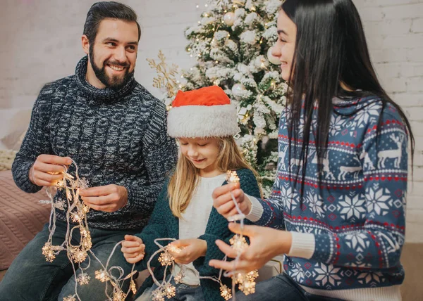 Schöne junge Familie genießt ihre gemeinsame Weihnachtszeit, schmückt den Weihnachtsbaum, arrangiert die Weihnachtsbeleuchtung und hat Spaß — Stockfoto