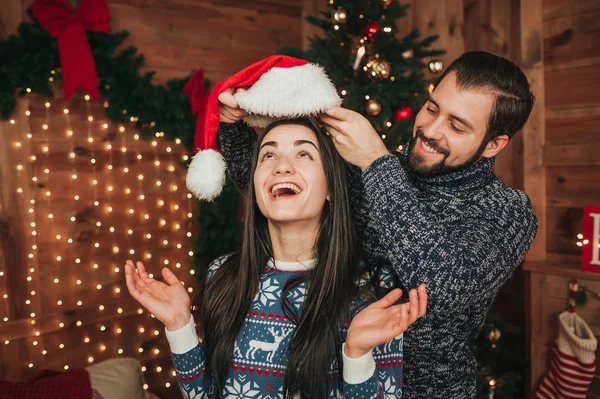 Feliz Navidad y Feliz Año Nuevo. Pareja joven celebrando vacaciones en casa. El hombre viste un sombrero de Navidad en una mujer — Foto de Stock