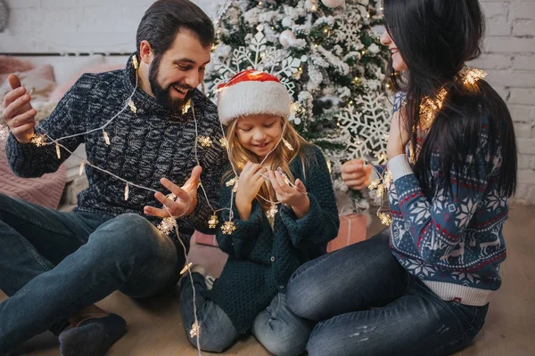 Hermosa familia joven disfrutando de sus vacaciones juntos, decorando el árbol de Navidad, organizando las luces de Navidad y divirtiéndose — Foto de Stock