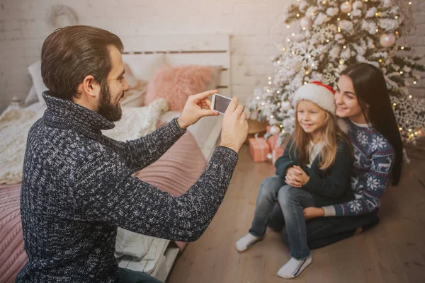 Familia joven sonriente en ambiente navideño haciendo foto con teléfono inteligente . — Foto de Stock