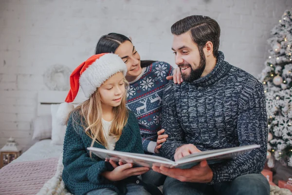 Merry Christmas and Happy Holidays Cheerful mom, dad and her cute daughter girl reading a book. Parent and little child having fun near Christmas tree indoors. Morning Xmas. Portrait family close up — Stock Photo, Image