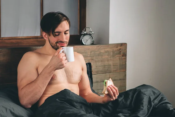 Красивый улыбающийся молодой голый мужчина держит чашку кофе и смотрит в сторону, сидя на кровати — стоковое фото