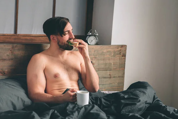 Красивый улыбающийся молодой голый мужчина держит чашку кофе и смотрит в сторону, сидя на кровати — стоковое фото