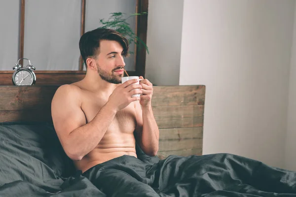 Красивий усміхнений молодий голий чоловік тримає чашку кави і дивиться геть, сидячи на ліжку — стокове фото