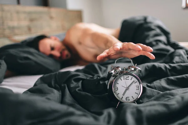 Ώρα να ξυπνήσουν. Κουρασμένος άνθρωπος στο κρεβάτι δεν είναι ευχαριστημένοι. Ώριμη τύπος κρατώντας ρολόι συναγερμών ενώ ελέγχετε ώρα για δουλειά — Φωτογραφία Αρχείου