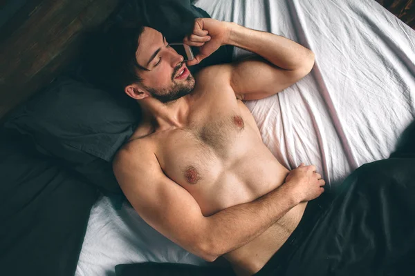 Pemuda ceria berbicara dengan ponsel di kamar tidur. Ganteng sporty pemuda bercelana dalam berbaring di tempat tidur — Stok Foto