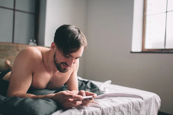 Cheerful young man speaking by cellphone in bedroom. Handsome sporty young guy in underwear is lying on bed