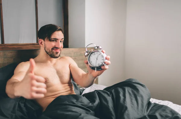 Прокинувшись хлопець. Чоловік тримає будильник бородатий голий людиною показ великі пальці на ліжку — стокове фото