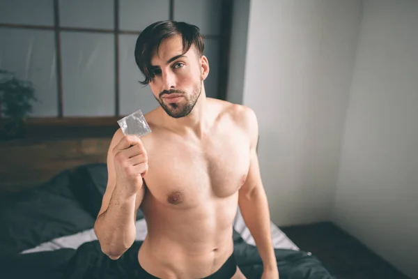 Emniyet seks konsepti. Koyu saçlı yakışıklı gülümseyen adam prezervatif yatakta elinde tutan bir çıplak kırpılmış görüntü sakallı — Stok fotoğraf