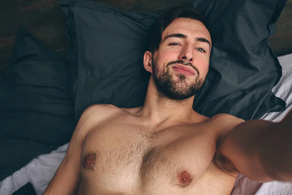 Nu barbudo escuro-cabelo bonito homem sem camisa no branco cama tomando um selfie — Fotografia de Stock