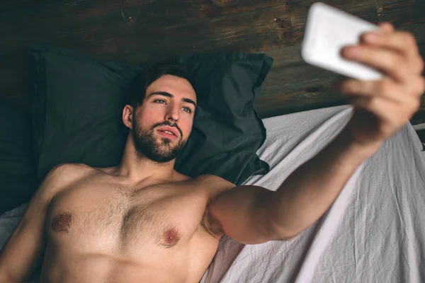 Голий бородатий темношерстий красивий чоловік без сорочки в білому ліжку приймає селфі — стокове фото