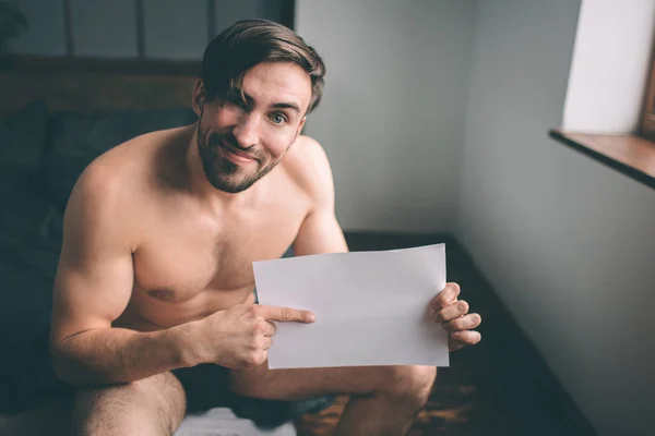 Desnudo barbudo de pelo oscuro guapo que sostiene una hoja de papel vacía en la cama. Interesante para el hogar — Foto de Stock