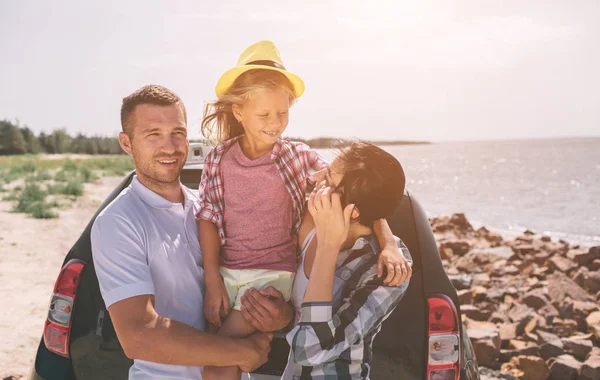 Jeune couple debout près de la botte de voiture ouverte avec valises et sacs. Papa, maman et fille voyagent par la mer, l'océan ou la rivière. Promenade estivale en automobile . — Photo