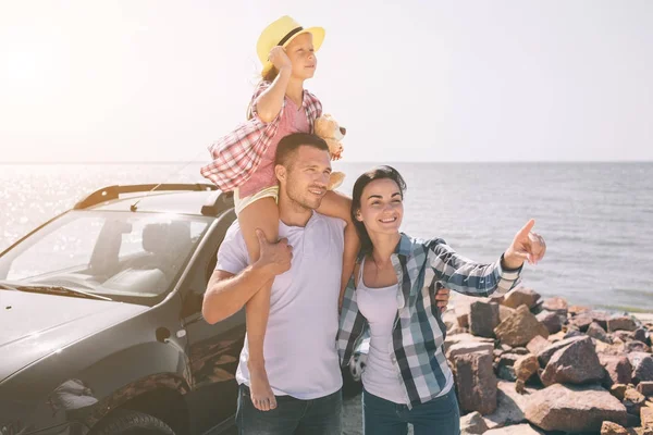 Familia feliz de pie cerca de un coche en la playa. Familia feliz en un viaje por carretera en su coche. Papá, mamá e hija están viajando por el mar o el océano o el río. Paseo de verano en automóvil — Foto de Stock