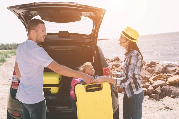 Young couple standing near the opened car boot with suitcases and bags. Dad, mom and daughter are traveling by the sea or the ocean or the river. Summer ride by automobile. — Stock Photo, Image