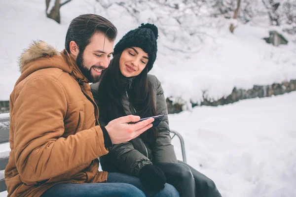 冬の幸せな若いカップル。屋外の家族。男と女の上方への検索と笑っています。愛、喜び、シーズン、冬の公園を歩いて人々。ベンチに座っています。スマート フォンを保持します。. — ストック写真