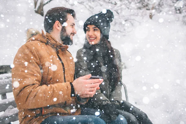 快乐的年轻夫妇在冬天。家庭户外。男人和女人看向上和笑。爱, 乐趣, 季节和人-漫步在冬天公园。下雪了, 他们在拥抱。坐在长凳上. — 图库照片
