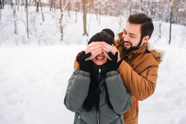 Счастливая молодая пара зимой. Семья на открытом воздухе. мужчина и женщина смотрят вверх и смеются. Любовь, веселье, сезон и люди - прогулки в зимнем парке. Он закрыл ей глаза руками. — стоковое фото