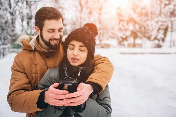 Glückliches junges Paar im Winter. Familie im Freien. Mann und Frau blicken nach oben und lachen. Liebe, Spaß, Jahreszeit und Menschen - Wandern im Winterpark. Es schneit, sie umarmen sich. sie trinken Kaffee. — Stockfoto