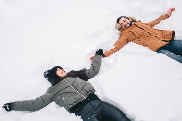 冬の幸せな若いカップル。屋外の家族。男と女の上方への検索と笑っています。愛、喜び、シーズン、冬の公園を歩いて人々。雪の天使たちが楽しい時を過すを作る新鮮な雪の中で横になっています。 — ストック写真
