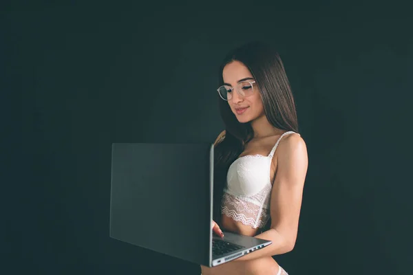 Donna usa il computer portatile. Giovane donna con lunghi capelli neri che indossa occhiali si trova isolata su sfondo nero. La ragazza ha una figura sportiva, è vestita con biancheria intima bianca — Foto Stock